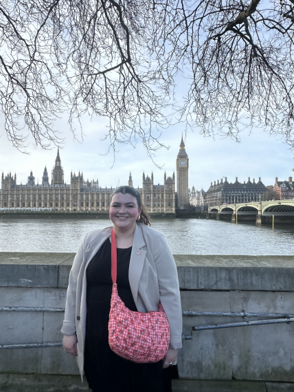Emily Wheaton smiling in front of the River Thames in London with the Palace of Westminster in the background.